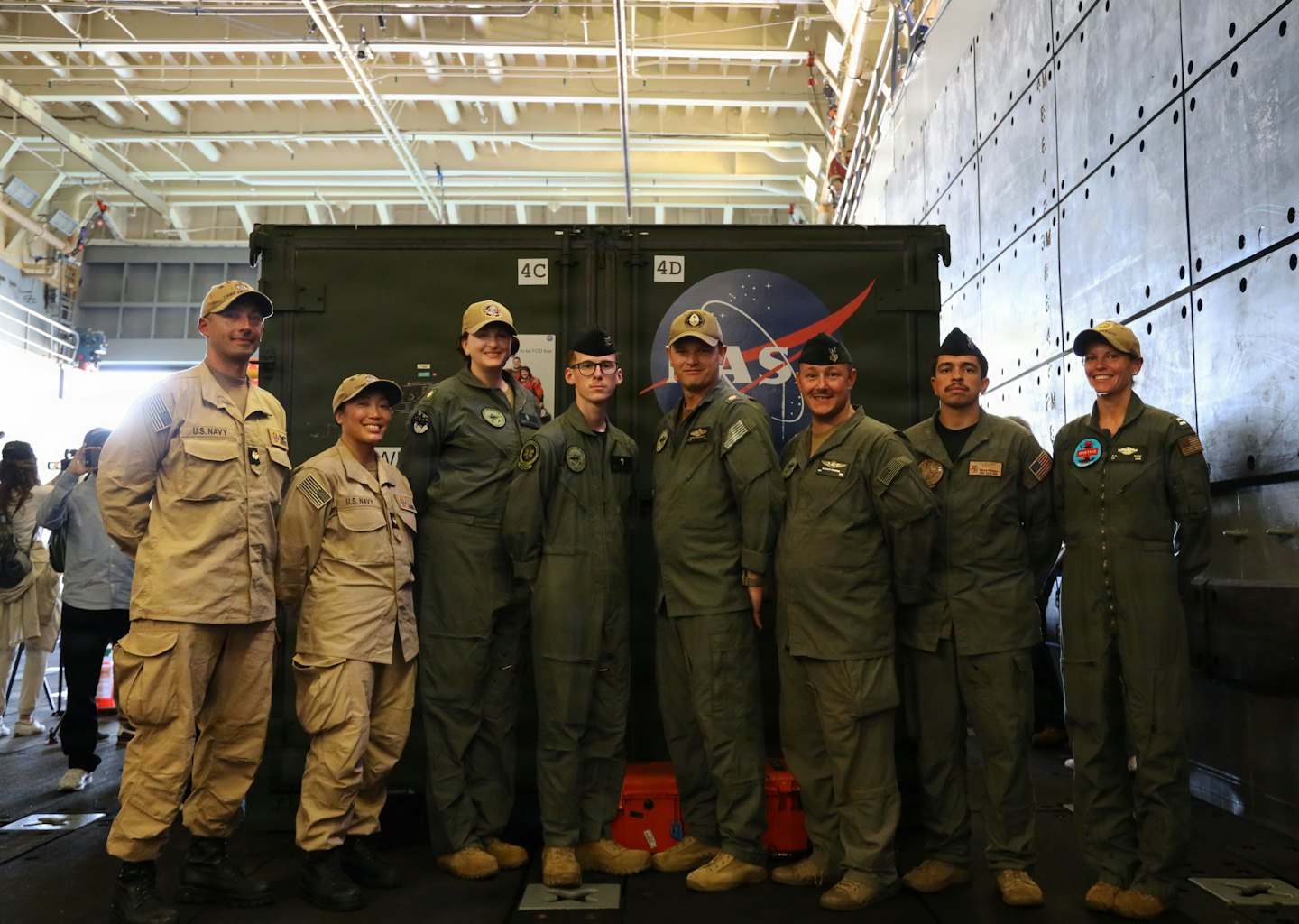Eight military personnel in uniforms stand before a NASA container.